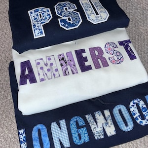 May include: Three hooded sweatshirts stacked on top of each other. The top sweatshirt is navy blue with "PSU" in white and blue patterned letters. The middle sweatshirt is white with "AMHERST" in purple patterned letters. The bottom sweatshirt is navy blue with "LONGWOOD" in blue patterned letters.