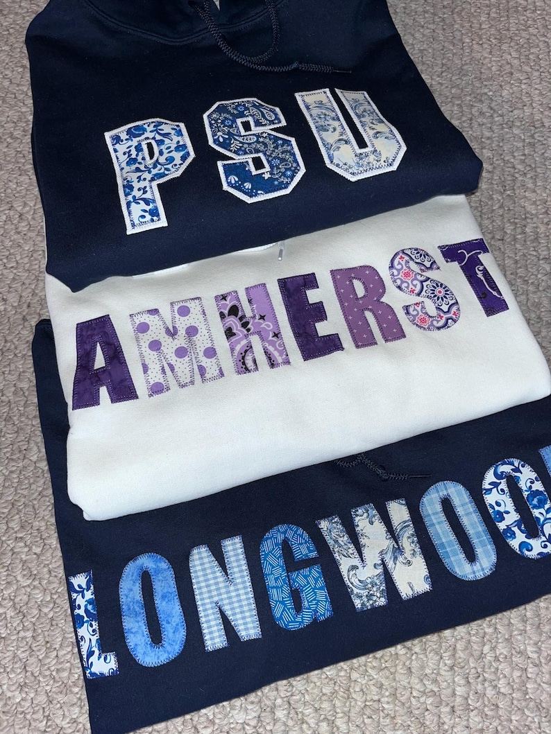 May include: Three hooded sweatshirts stacked on top of each other. The top sweatshirt is navy blue with "PSU" in white and blue patterned letters. The middle sweatshirt is white with "AMHERST" in purple patterned letters. The bottom sweatshirt is navy blue with "LONGWOOD" in blue patterned letters.