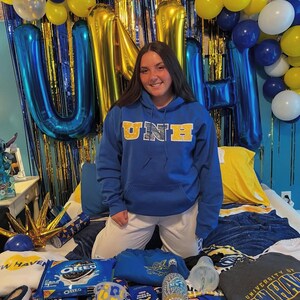 May include: A blue hoodie with "UNE" in gold lettering is worn by a person kneeling on a bed. The bed is covered with gifts, including snacks, a Starbucks cup, and a sweatshirt. Blue and gold balloons and streamers decorate the background.