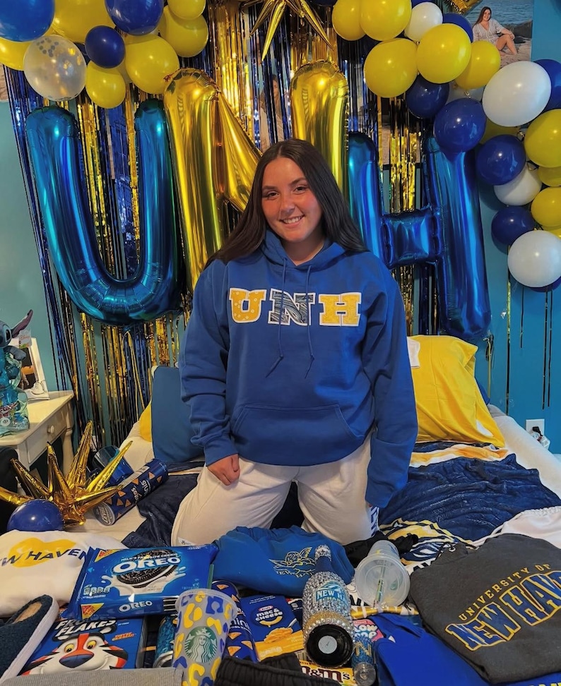 May include: A blue hoodie with "UNE" in gold lettering is worn by a person kneeling on a bed. The bed is covered with gifts, including snacks, a Starbucks cup, and a sweatshirt. Blue and gold balloons and streamers decorate the background.