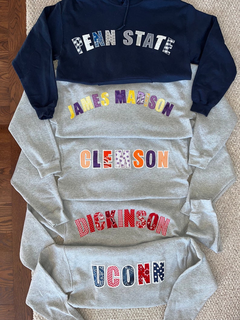 May include: A stack of five sweatshirts, each with a different college name appliqued across the chest. The top sweatshirt is navy blue with "PENN STATE" in patchwork letters. The others are gray, with "JAMES MADISON", "CLEMSON", "DICKINSON", and "UCONN".