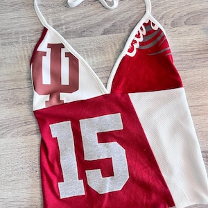 May include: A halter top with a white tie-neck and a patchwork design. The top features a red panel with the number 15 in gray, a white panel with a textured pattern, and a panel with the Indiana University logo in maroon.