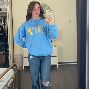 May include: A light blue hoodie with the word "PITT" in yellow and blue letters. The person in the image is wearing blue jeans with a distressed knee. The person is holding a phone and taking a mirror selfie.