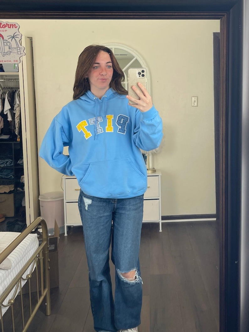 May include: A light blue hoodie with the word "PITT" in yellow and blue letters. The person in the image is wearing blue jeans with a distressed knee. The person is holding a phone and taking a mirror selfie.