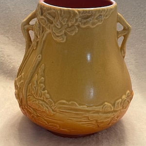 May include: A ceramic vase with a yellow-orange gradient exterior and a red interior. The vase features two handles and embossed floral and landscape designs. The base is a darker shade of orange.