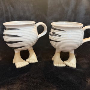 May include: Two white ceramic mugs with black stripe accents and beige feet. The mugs have a round body, a handle, and are supported by two beige feet. The mugs are on a dark background.