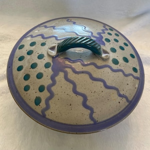 May include: A ceramic pot lid with a teal handle and rim. The lid is speckled with teal dots and wavy purple lines. The rim is a solid purple color. The handle is a twisted design.