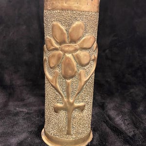 May include: A vintage brass vase with a textured surface and a large floral design. The vase has a scalloped rim and a cross-like design at the base of the flower. The brass has a warm, aged patina.