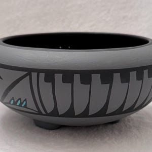 以下が含まれることがあります： 丸みを帯びた形状で3つの小さな脚が付いたマットグレーの陶器製ボウル。外側には、繰り返される垂直の黒い形状と、3つの小さな明るいターコイズブルーのアクセントが入った幾何学的な三角形のパターンで装飾された幅の広い黒い帯が施されています。