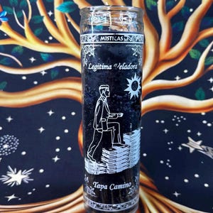 May include: A black glass candle with white text and illustrations. The candle features a man climbing steps, a sun, and the words "Misticas", "Legitima Veladora", and "Tapa Camino". The background is a tree design.