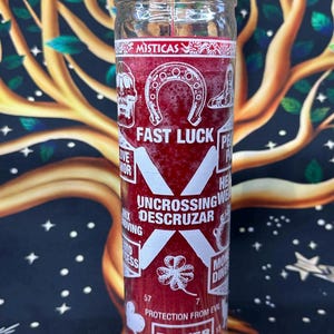 May include: A red glass candle with white text and symbols, including a horseshoe, skull, and four-leaf clover. The candle reads "Fast Luck" and "Uncrossing/Descruzar", with additional text indicating protection and power.