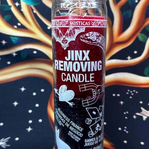 May include: A glass jar candle with a red and black label. The label reads "Jinx Removing Candle" with illustrations of a bat, snake, and other symbols. Additional text includes "Business Finance" and "Helping Hand for Success."