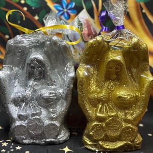May include: Two decorative candles, one silver and one gold, each shaped like a hooded figure. The candles are wrapped in clear cellophane and tied with a yellow and purple ribbon. The candles have a detailed design.