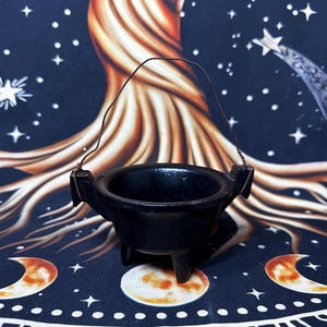 May include: A small, black, three-legged cast iron cauldron featuring a thin, arched wire handle attached to two side lugs. The vessel has a wide, open circular top and a smooth, dark finish, designed for holding small items or decorative use.