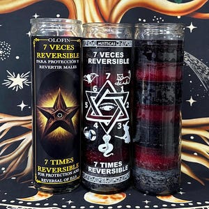 May include: Three tall cylindrical glass candles with layered black and red wax. The left candle features a star graphic and text reading 7 VECES REVERSIBLE. The center candle displays a hexagram with an eye and the text 7 VECES REVERSIBLE.