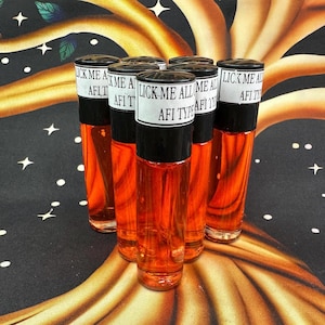 May include: A group of six glass bottles with black caps and white labels. The bottles are filled with a bright orange liquid. The labels read "LICK ME ALL AFI TYPE". The bottles are arranged on a patterned surface.