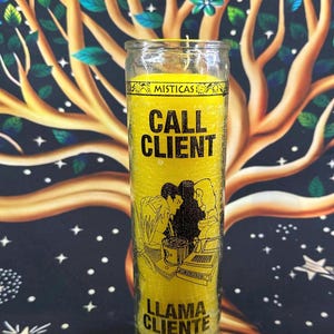 May include: A yellow candle with the words "CALL CLIENT" in bold black letters. The candle has an illustration of people working on a laptop and a phone. The bottom of the candle reads "LLAMA CLIENTE". The candle is in a clear glass jar.