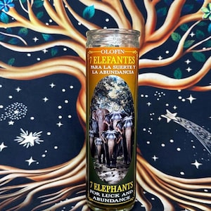 May include: A cylindrical glass candle featuring a label with an image of seven elephants. The text reads OLOFIN 7 ELEFANTES PARA LA SUERTE Y LA ABUNDANCIA and 7 ELEPHANTS FOR LUCK AND ABUNDANCE. The label has a gold and brown color scheme.