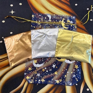 May include: An overhead shot of three small, shiny pouches in gold, silver, and bronze, arranged on a celestial-themed fabric. A blue mesh bag with gold stars, moons, and a drawstring is also visible.