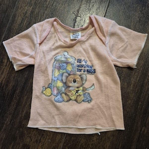 May include: A light peach-colored baby t-shirt with short sleeves. The shirt features a graphic of a koala bear holding a sign that says "I'd do anything for a KISS" and a jar filled with colorful candies.