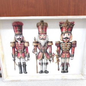 May include: A rectangular wooden sign with a white frame features three detailed illustrations of nutcrackers in red, gold, and white uniforms. Each nutcracker has a unique hat and holds a staff or weapon. The sign is a festive holiday decoration.