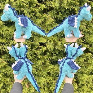 May include: Four views of a crocheted dragon in shades of blue, white, and navy. The dragon has a long tail, wings, and horns. The dragon is held up by a hand.