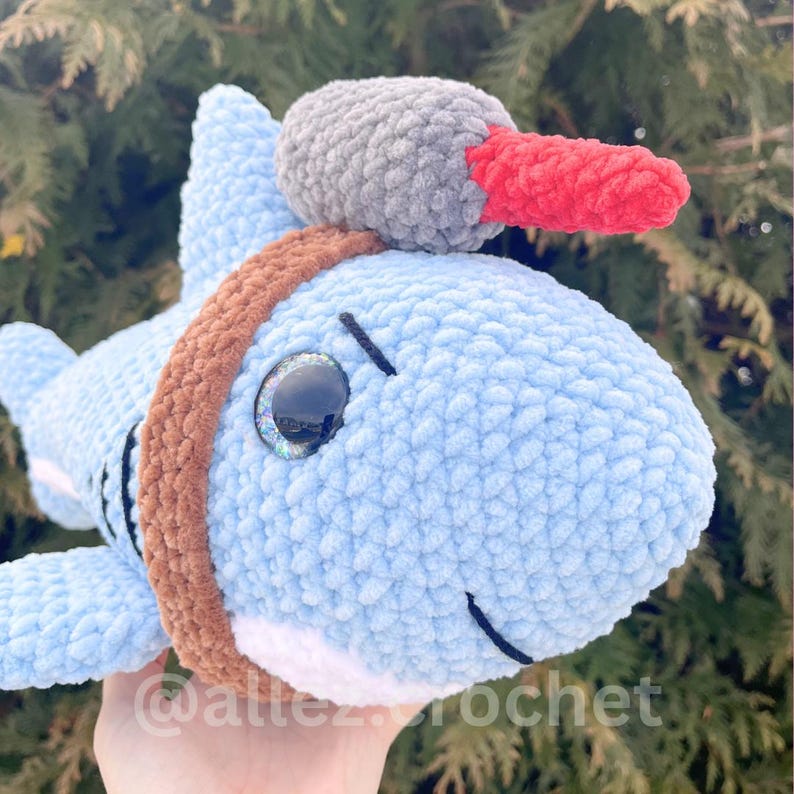 Puede incluir: Un peluche de tibur&oacute;n de ganchillo azul claro con una banda marr&oacute;n y un sombrero gris y rojo. El tibur&oacute;n tiene ojos grandes y brillantes y una barriga blanca. El peluche es suave y tierno, ideal para ni&ntilde;os.