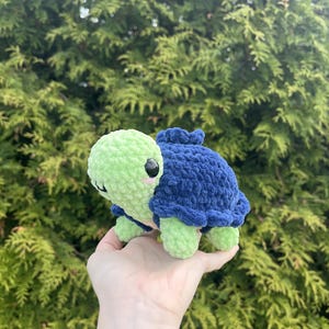 May include: A small, handmade crochet turtle plush toy featuring a light green head and limbs with a dark blue textured shell. The turtle has black plastic safety eyes and small pink embroidered cheeks on its round, soft-textured face.