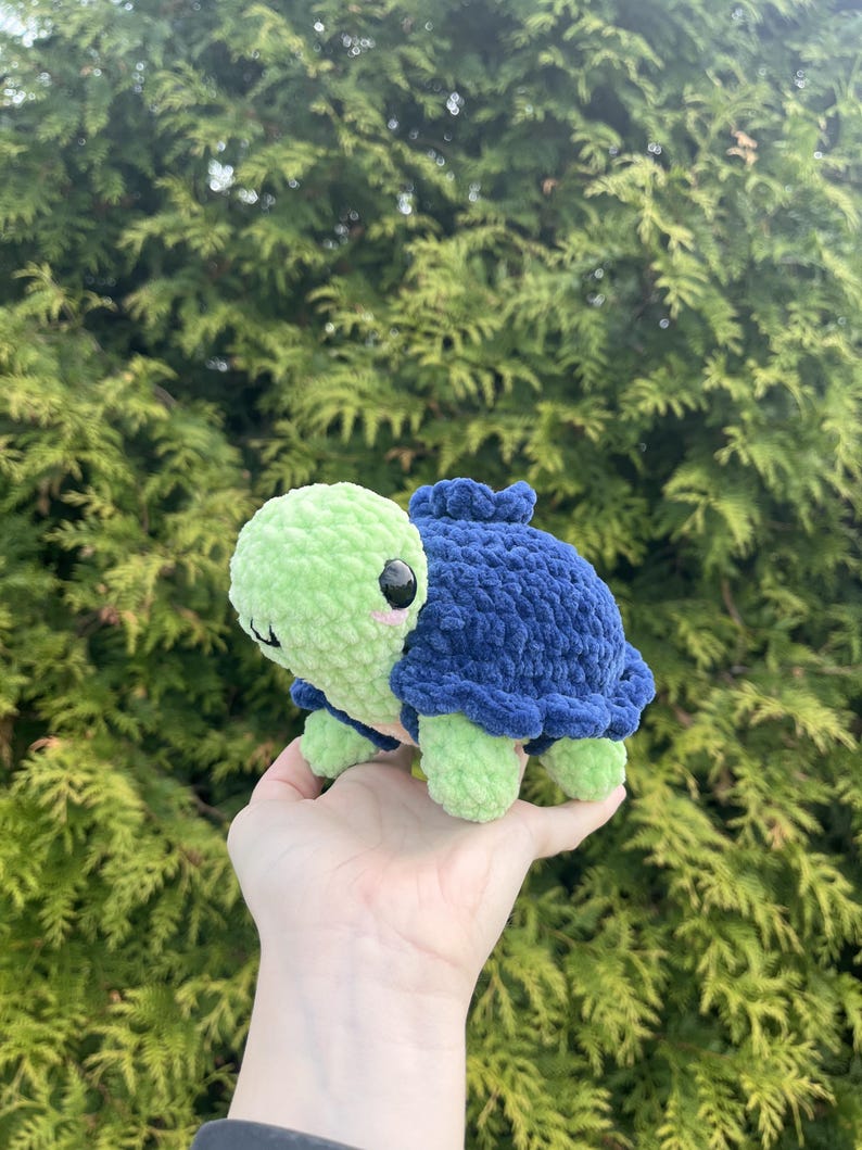 May include: A small, handmade crochet turtle plush toy featuring a light green head and limbs with a dark blue textured shell. The turtle has black plastic safety eyes and small pink embroidered cheeks on its round, soft-textured face.
