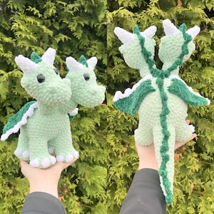 May include: Two-headed crochet dragon in light green with white and dark green accents. The plush toy has white horns, green wings, and a long tail. The dragon is handmade and has a soft, textured appearance, perfect for a child's toy or room decor.
