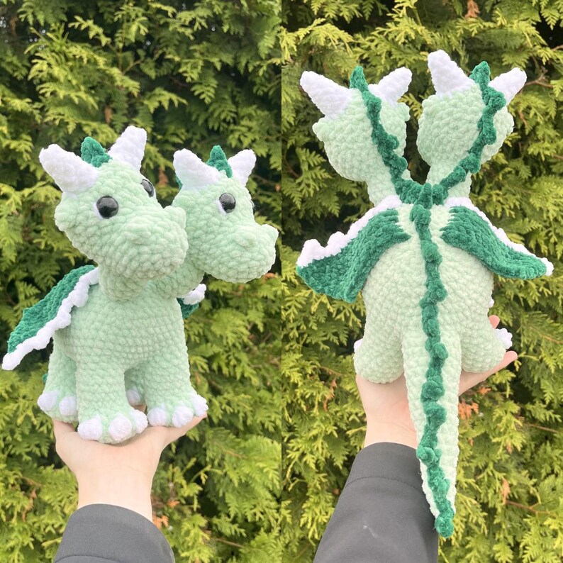 May include: Two-headed crochet dragon in light green with white and dark green accents. The plush toy has white horns, green wings, and a long tail. The dragon is handmade and has a soft, textured appearance, perfect for a child's toy or room decor.