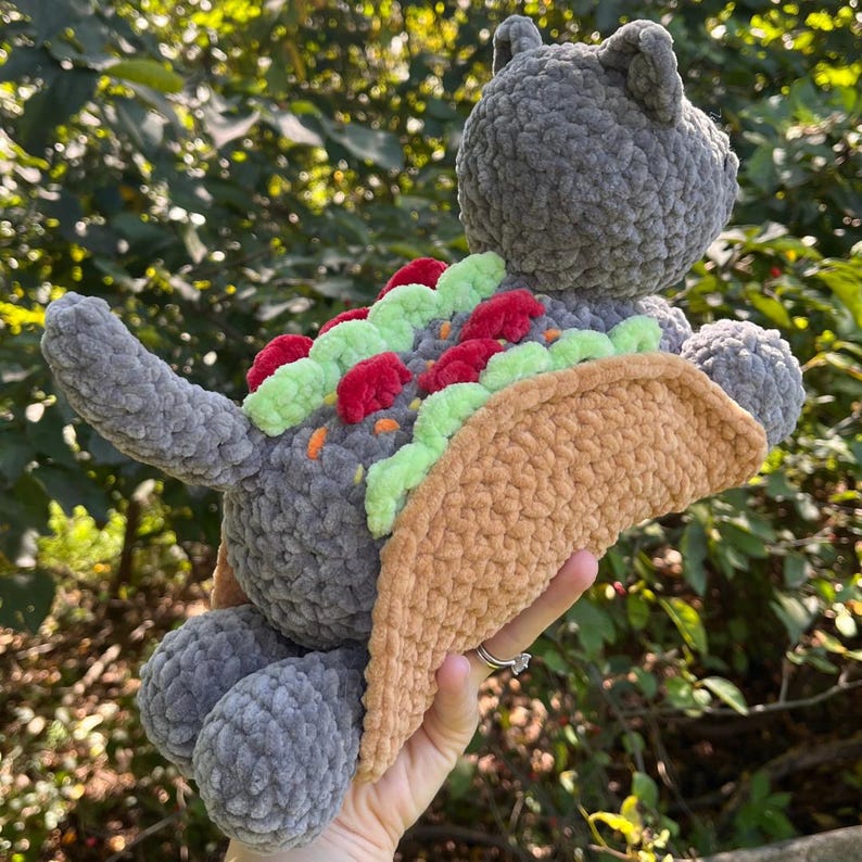 Puede incluir: Un juguete de peluche gris con forma de gato con un cuerpo de concha de taco. El taco est&aacute; relleno de ingredientes de peluche verdes, rojos y naranjas. El gato tiene cola, orejas y patas. El juguete es hecho a mano y suave.