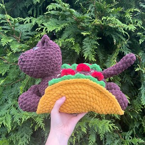 Puede incluir: Un juguete de dinosaurio de peluche morado con un gran taco amarillo. El taco est&aacute; relleno de lechuga verde y tomates rojos. El dinosaurio est&aacute; hecho de un material suave y texturizado.
