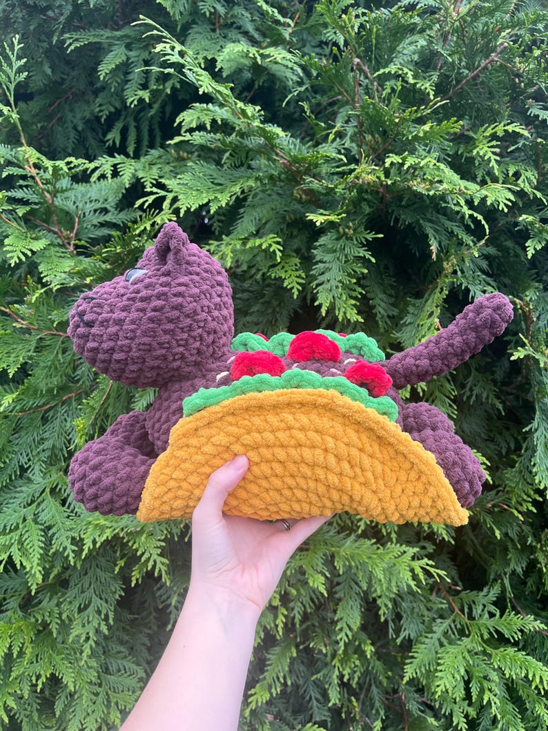 Puede incluir: Un juguete de dinosaurio de peluche morado con un gran taco amarillo. El taco est&aacute; relleno de lechuga verde y tomates rojos. El dinosaurio est&aacute; hecho de un material suave y texturizado.