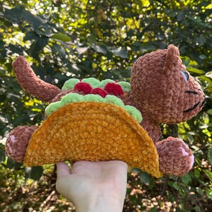 Puede incluir: Un juguete de peluche marr&oacute;n con forma de oso, con un cuerpo de concha de taco. El taco est&aacute; relleno de lechuga verde y tomates rojos. El oso tiene una cara sonriente y sostiene el taco. El juguete est&aacute; hecho de un material suave y texturizado.