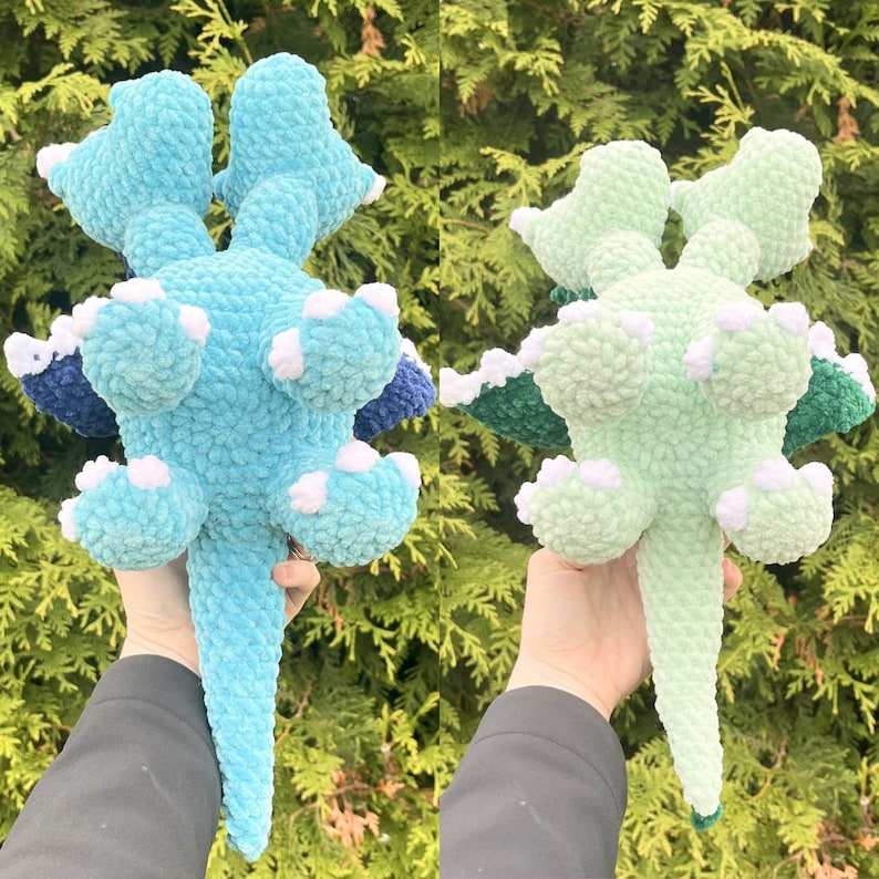 May include: Two plush, handmade crochet dinosaurs in the image. One is light blue with white and navy accents, the other is light green with white and dark green accents. The dinosaurs are held up against a backdrop of green foliage.