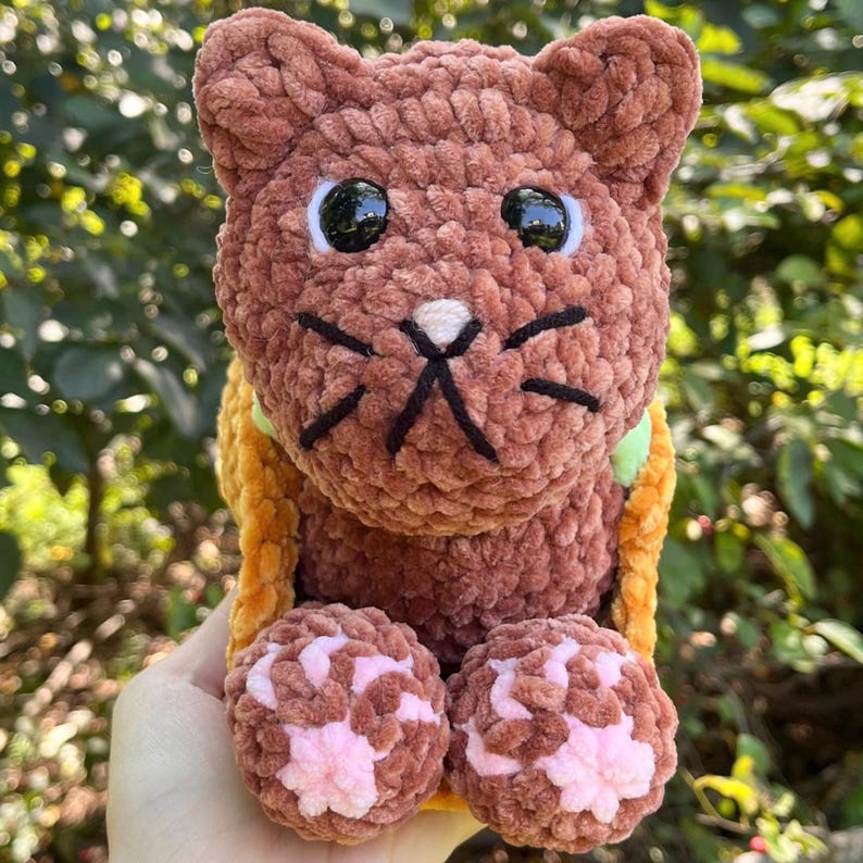 Puede incluir: Un peluche de gato de ganchillo marr&oacute;n hecho a mano con bigotes bordados negros y ojos grandes negros. El gato tiene patas rosas y marrones y lleva un collar amarillo y verde. El peluche es suave y tierno.