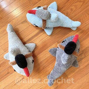 Puede incluir: Tres peluches de tibur&oacute;n de ganchillo en azul claro, gris y gris azulado. Cada tibur&oacute;n tiene una h&eacute;lice, una banda marr&oacute;n y un detalle rojo y negro. Los juguetes est&aacute;n colocados sobre una superficie de madera.