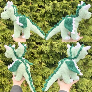 May include: A crocheted dragon toy in shades of light green and dark green. The dragon has white accents on its wings, horns, and feet. The toy is soft and plush, with a detailed design, and is displayed against a backdrop of green foliage.