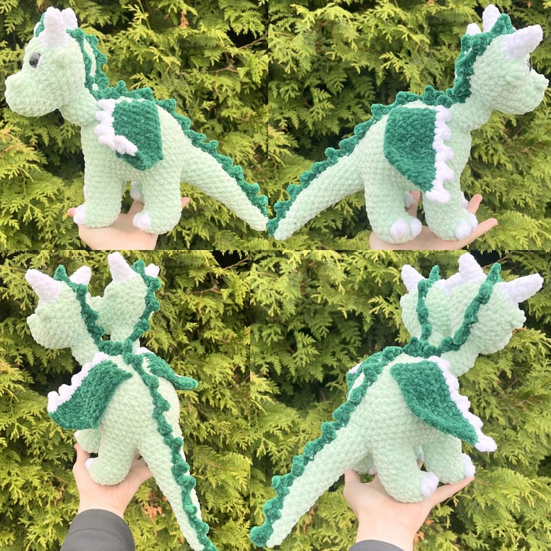 May include: A crocheted dragon toy in shades of light green and dark green. The dragon has white accents on its wings, horns, and feet. The toy is soft and plush, with a detailed design, and is displayed against a backdrop of green foliage.