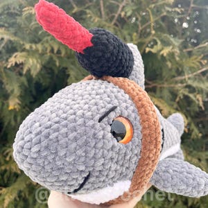 Puede incluir: Un peluche de tibur&oacute;n gris tejido a mano con un cuerno rojo y negro. El tibur&oacute;n tiene un collar marr&oacute;n, ojos naranjas y una barriga blanca. El peluche es suave y tierno, perfecto para ni&ntilde;os o como objeto decorativo.