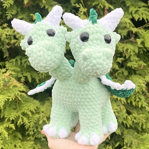 May include: A handmade, two-headed crochet dragon in soft green yarn. The dragon features white horns, accents, and claws, with black safety eyes. The wings are green and white. The plush toy is a whimsical, cuddly creature.