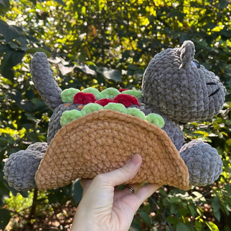 Puede incluir: Un peluche gris con forma de gato tejido con un cuerpo de concha de taco. El taco est&aacute; relleno de lechuga verde, tomates rojos y queso naranja. El gato tiene cola, orejas y una cara sonriente.