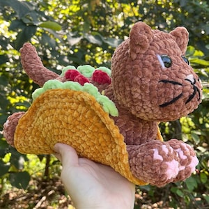 Puede incluir: Un peluche marr&oacute;n con forma de gato, con una concha de taco amarillo-naranja alrededor del cuerpo. El taco tiene ingredientes verdes y rojos. El gato tiene bigotes negros y almohadillas rosas.