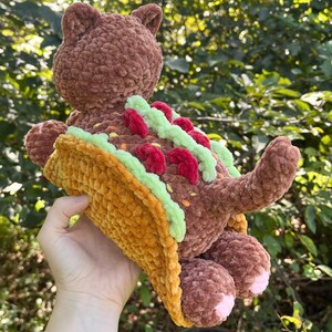 Puede incluir: Un peluche de oso marr&oacute;n tejido a ganchillo con un taco. El taco es amarillo, verde y rojo, y el oso tiene detalles rosas en las patas. El oso est&aacute; sobre un fondo de follaje verde.