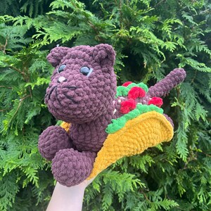 Puede incluir: Una criatura hecha a mano, de felpa, morada, con forma de gato, con grandes ojos negros, sosteniendo un taco amarillo con lechuga verde y aderezos de tomate rojo. El peluche est&aacute; colocado sobre un fondo de follaje verde.