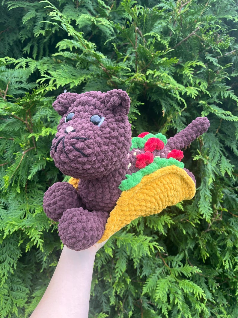 Puede incluir: Una criatura hecha a mano, de felpa, morada, con forma de gato, con grandes ojos negros, sosteniendo un taco amarillo con lechuga verde y aderezos de tomate rojo. El peluche est&aacute; colocado sobre un fondo de follaje verde.