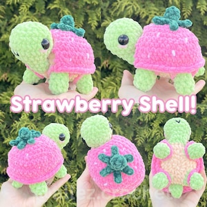 May include: A crochet turtle plush with a light green head and limbs, featuring a bright pink strawberry-shaped shell with white speckles and a dark green stem on top. The text Strawberry Shell! is displayed across the center of the image.