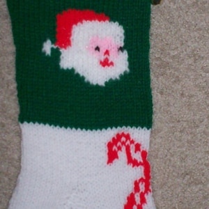 May include: Hand-knitted Christmas stocking with a red cuff and toe, white body, and green accents. The stocking features a Santa Claus face and a candy cane design. Includes a gold bell and a loop for hanging.