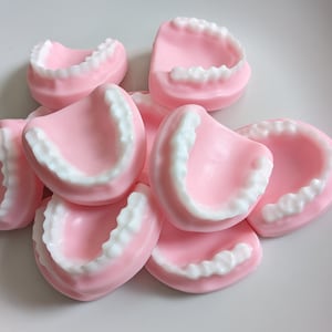 May include: A collection of pink, tooth-shaped objects with white, detailed teeth. The objects are arranged in a pile, with some overlapping. The background is a plain white surface. The objects appear to be novelty items or decorative pieces.